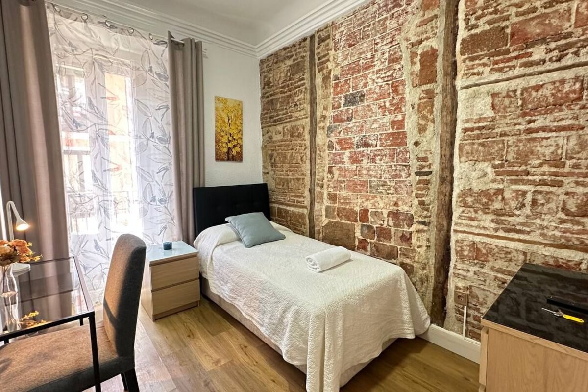 A bedroom with a brick wall, a bed with a white bedspread, a desk with a chair, and a window with curtains.