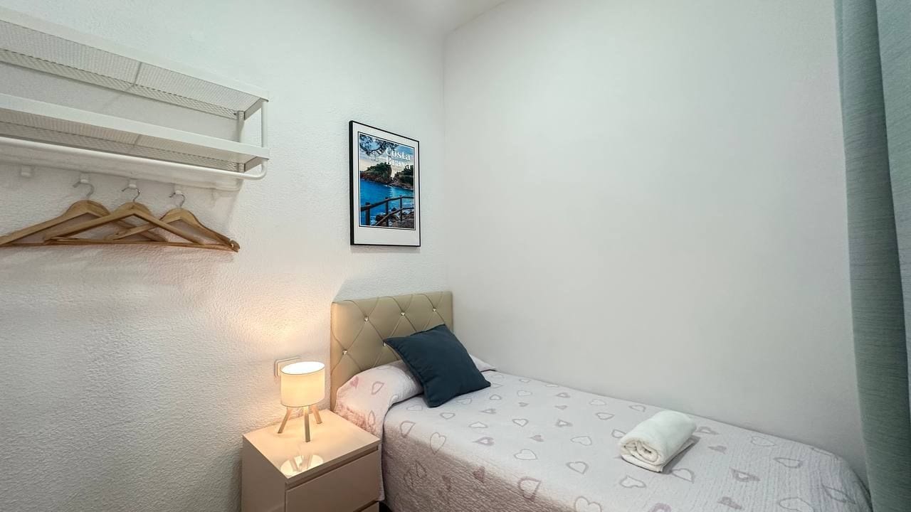 A small, neatly arranged bedroom with a bed, a bedside table with a lamp, a hanging coat rack, and a framed picture on the wall.