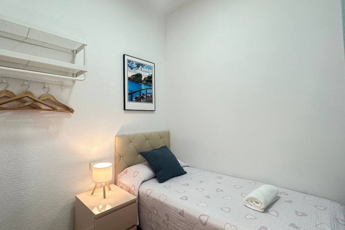 A small, neatly arranged bedroom with a bed, a bedside table with a lamp, a hanging coat rack, and a framed picture on the wall.