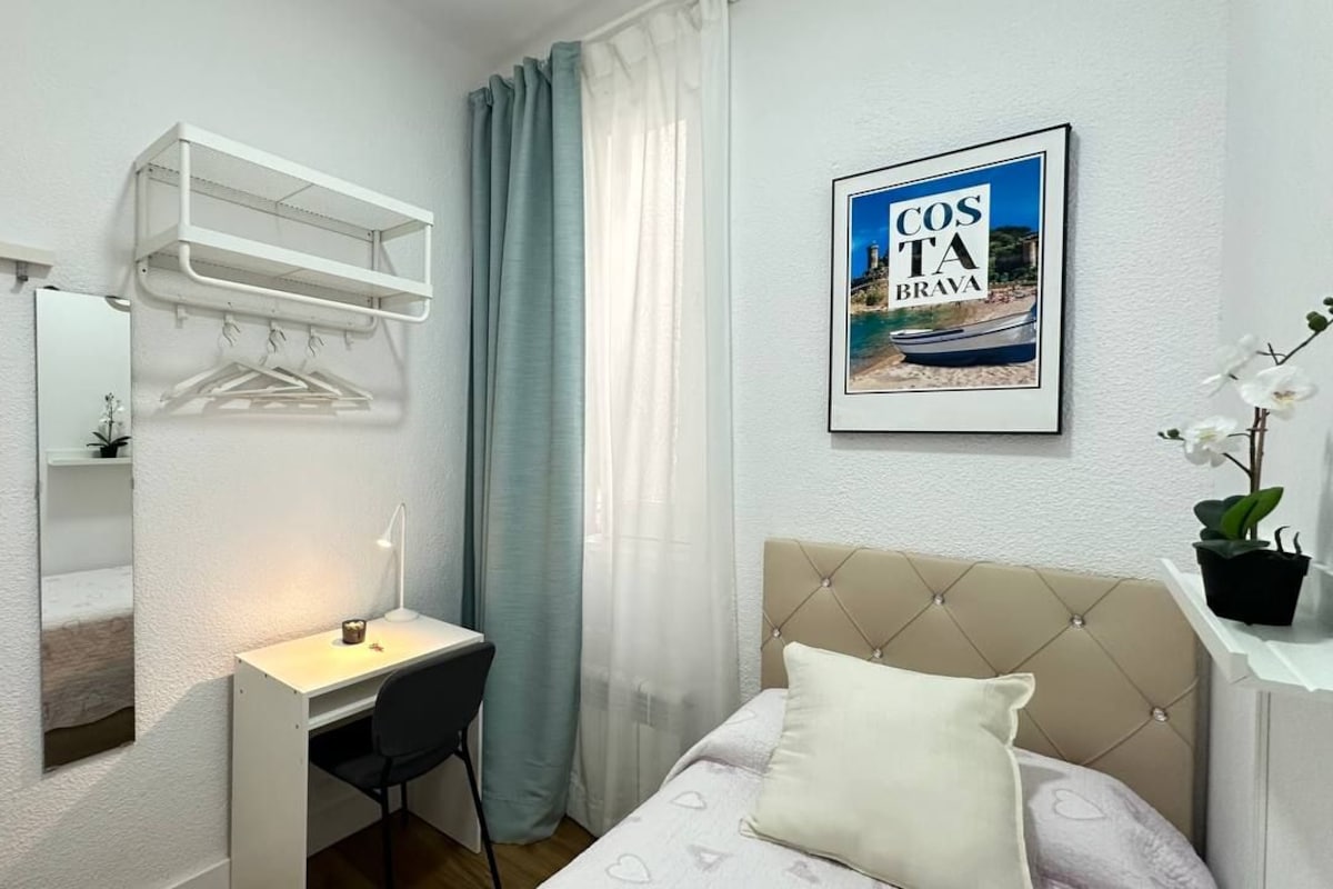 Habitación de hotel con cama, escritorio, silla, perchero, un cuadro de Costa da Praia y una planta en maceta.