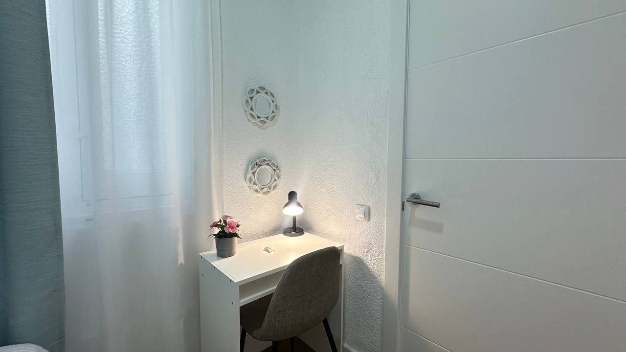 Een wit bureau met een lamp, een stoel en een potplant in een hoek van een kamer met witte muren en een deur.