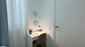 A white desk with a lamp, a chair, and a potted plant in a corner of a room with white walls and a door. - Habitaciones Malasaña (Madrid)