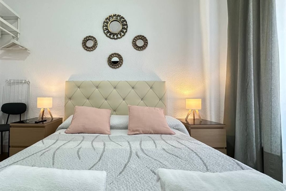 Un dormitorio ordenado con cabecera capitonada, dos lámparas de noche, un radiador, una silla y espejos decorativos en la pared.