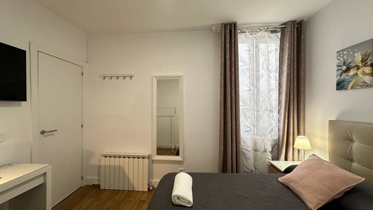 A hotel room with a bed, a bedside table, a mirror, a radiator, a TV, and a window with curtains.
