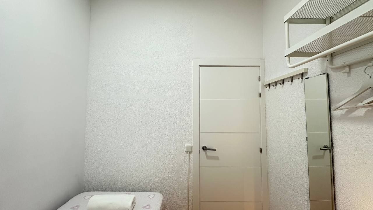 Een kleine kamer met witte wanden, een deur, een spiegel en een plank met hangende spullen.