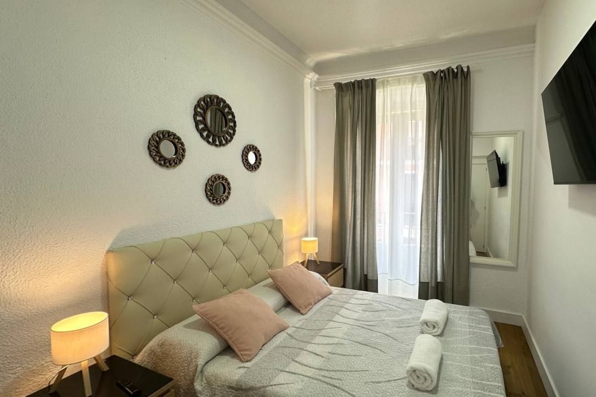 Habitación de hotel con cabecera capitonada, cama con colcha, dos lámparas de noche, televisor de pantalla plana en la pared y ventana con cortinas.