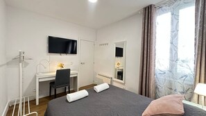 A bedroom with a bed, a desk, a chair, a TV, a mirror, and a window with curtains. - Habitaciones Malasaña (Madrid)