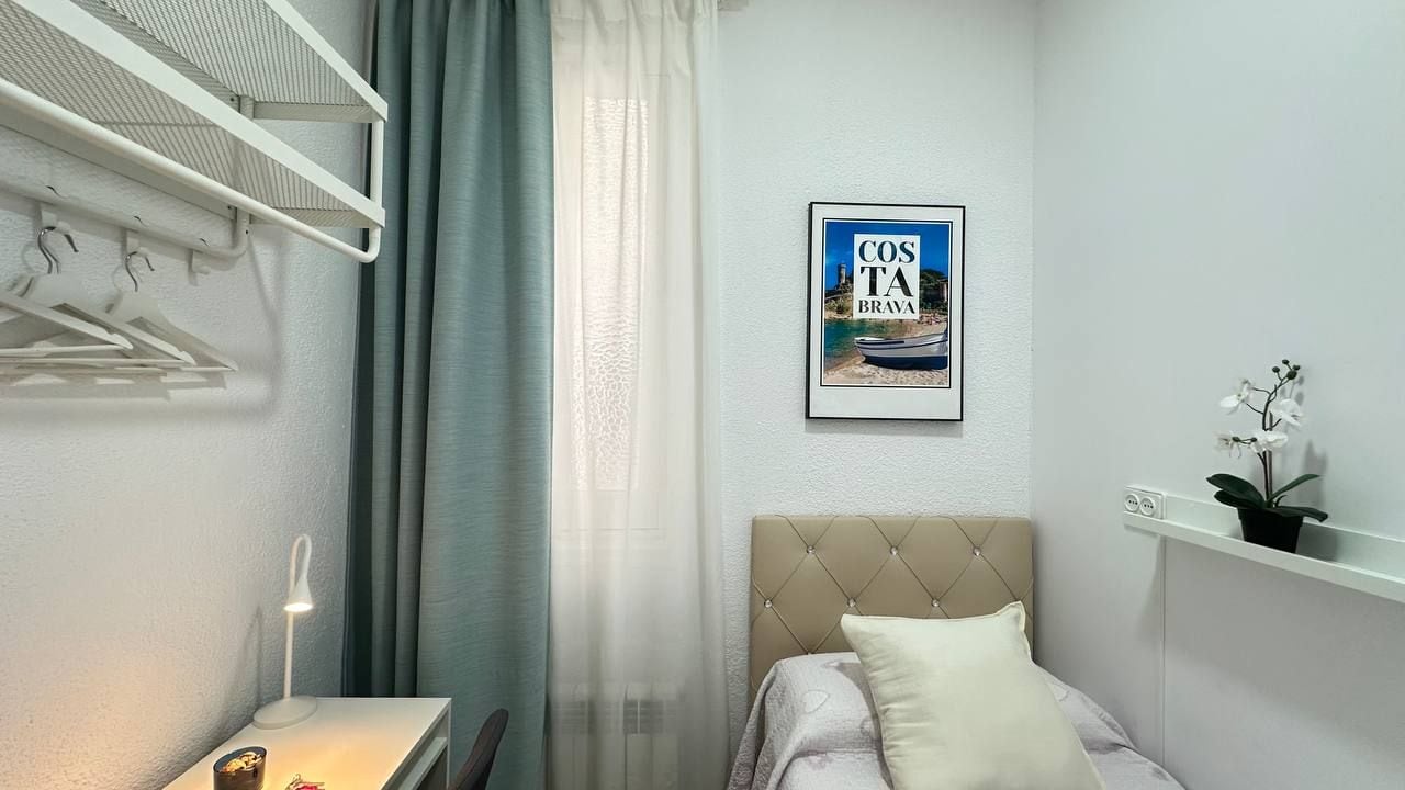 A bedroom with a bed, a bedside table, a lamp, a wall-mounted shelf with a plant, a framed picture, and a window with curtains.