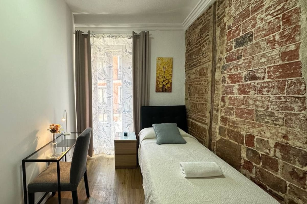 Una habitación pequeña y acogedora con pared de ladrillo, cama con almohadas, escritorio con silla y ventana con cortinas.