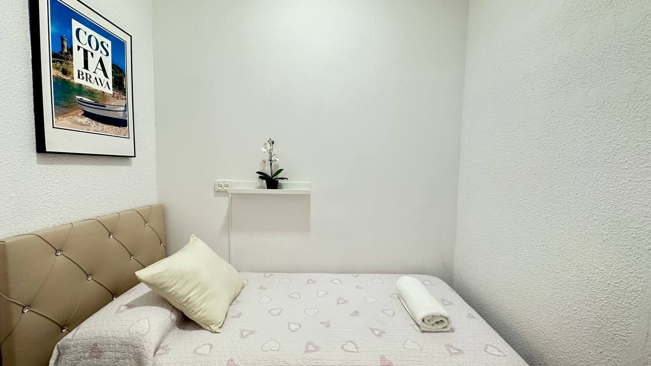 Een slaapkamer met een bed, een kussen, een opgerolde handdoek, een klein plankje met een plantje en een ingelijste foto aan de muur.