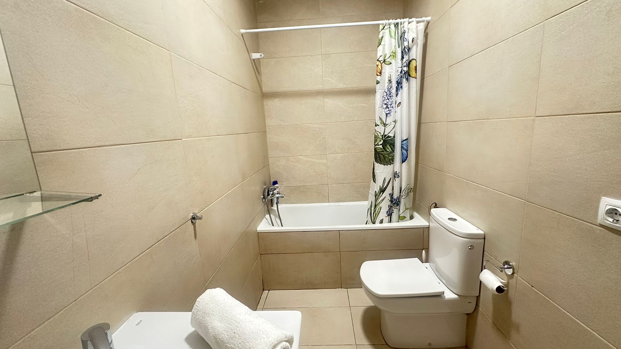 Un baño pequeño con inodoro, estante de vidrio, bañera con cortina de ducha y paredes con azulejos color beige.