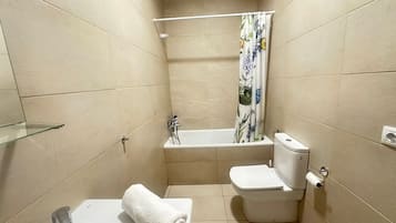 Un baño pequeño con inodoro, estante de vidrio, bañera con cortina de ducha y paredes con azulejos color beige.