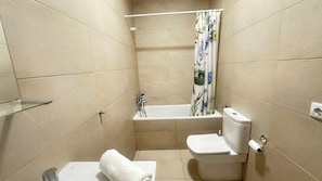 Un baño pequeño con inodoro, estante de vidrio, bañera con cortina de ducha y paredes con azulejos color beige.