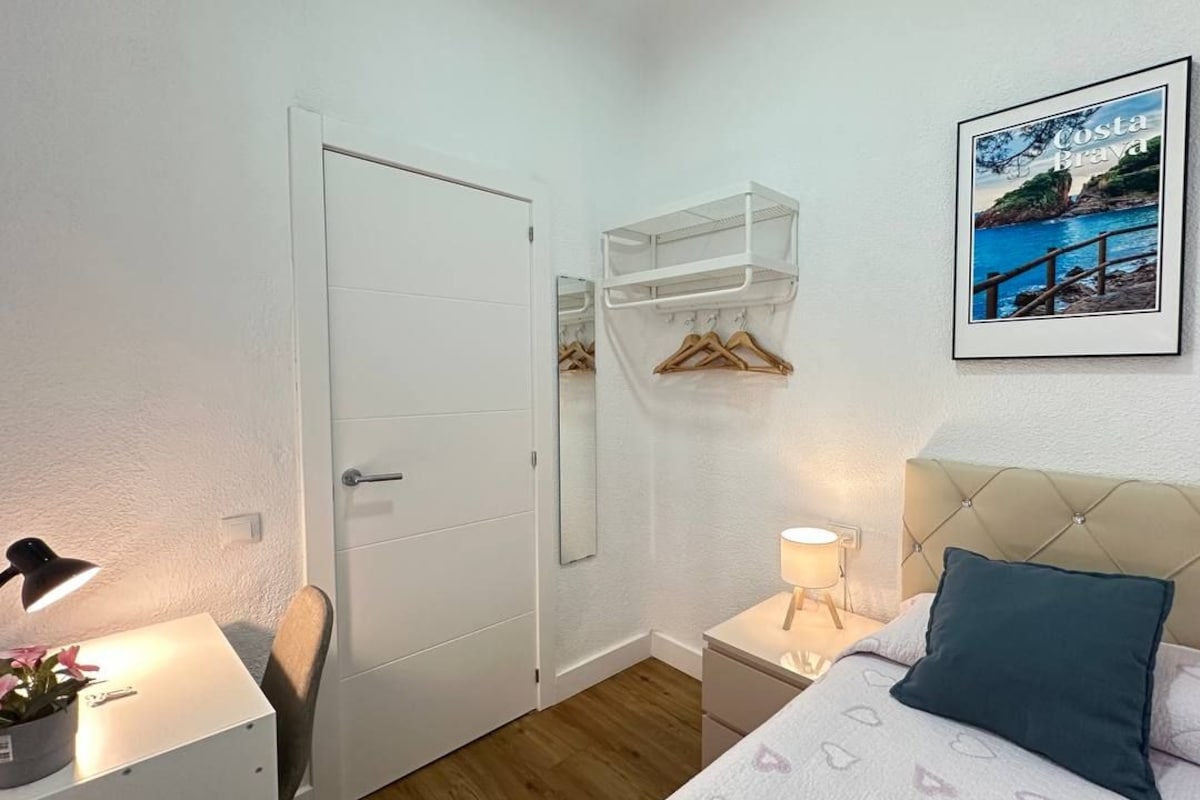 Un dormitorio con cama, mesita de noche, lámpara, silla, armario y un cuadro en la pared.