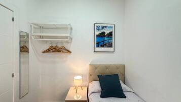 A bedroom with a bed, a bedside lamp, a hanging rack with clothes, a framed picture, and a mirror.