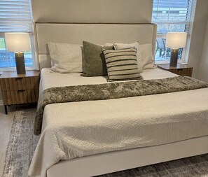 A neatly made bed with a headboard, two bedside tables with lamps, and a view of the outside through the window. - NEW 3BR Pool Home on Woodlands 10th Green, Walk to Eastport, 4 Seater Golf Cart (The Villages)
