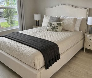 Cama com cabeceira branca, manta cinza e várias almofadas, incluindo uma com desenho de palmeira.
