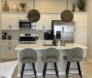 A modern kitchen with white cabinets, a stainless steel refrigerator, a microwave, and a gas stove. There are three bar stools with black legs. - NEW 3BR Pool Home on Woodlands 10th Green, Walk to Eastport, 4 Seater Golf Cart (The Villages)