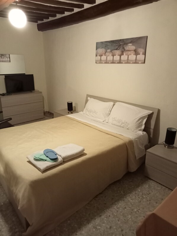 A bedroom with a bed, a nightstand, a lamp, a television on a dresser, and a painting on the wall.