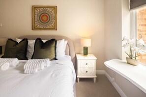 A neatly made bed with white linens and rolled towels, a bedside lamp, a decorative wall piece, and a window with white orchids. - 10 Mins Walk to Selfridges Marylebone Retreat (London)