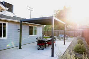 Ein überdachter Patio-Bereich mit Tisch und Stühlen, einer Pergola und Blick auf ein Wohnhaus.
