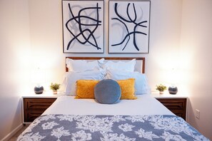 Een net opgemaakt bed met een bloemenprintdeedbedovertrek, twee nachtkastjes met decoratieve vazen, en abstracte kunst aan de muur.