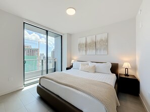 A modern bedroom with a large bed, a nightstand, a wall-mounted lamp, and a city view through a sliding glass door. - Luxury 2BR Apt| Water & Skyline Views | 29th Floor (Miami)