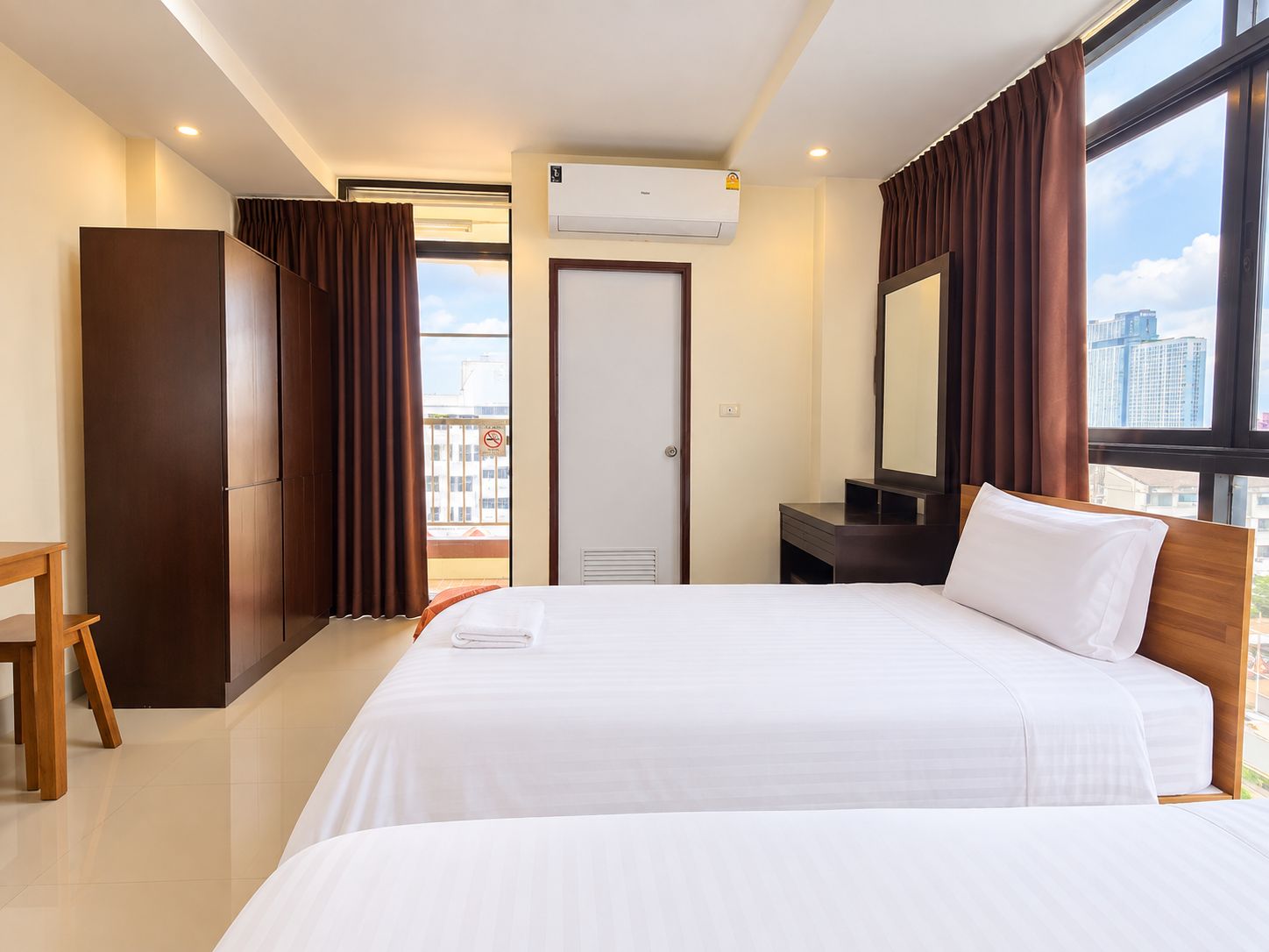 Quarto de hotel com cama, escrivaninha, guarda-roupa, espelho e janela com cortinas.