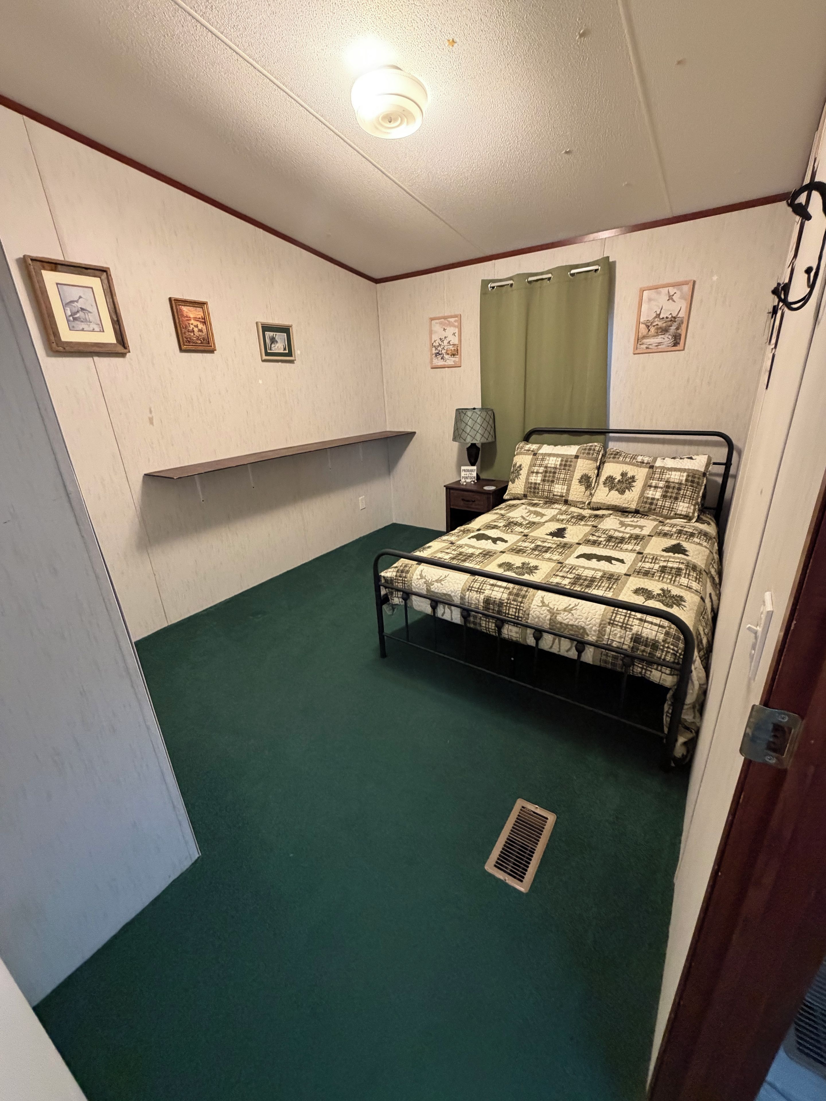 A bedroom with a bed, a nightstand, a wall-mounted shelf, and a window with a green curtain.