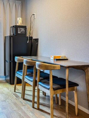 A compact room with a wooden dining table, two matching chairs, a black refrigerator, and a small kitchenette area with a microwave. - HANASTAY TAKE (Osaka)
