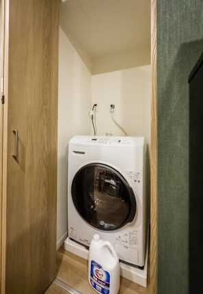 A compact, white front-loading washing machine in a small laundry room with a wooden door and a green wall. - HANASTAY TAKE (Osaka)