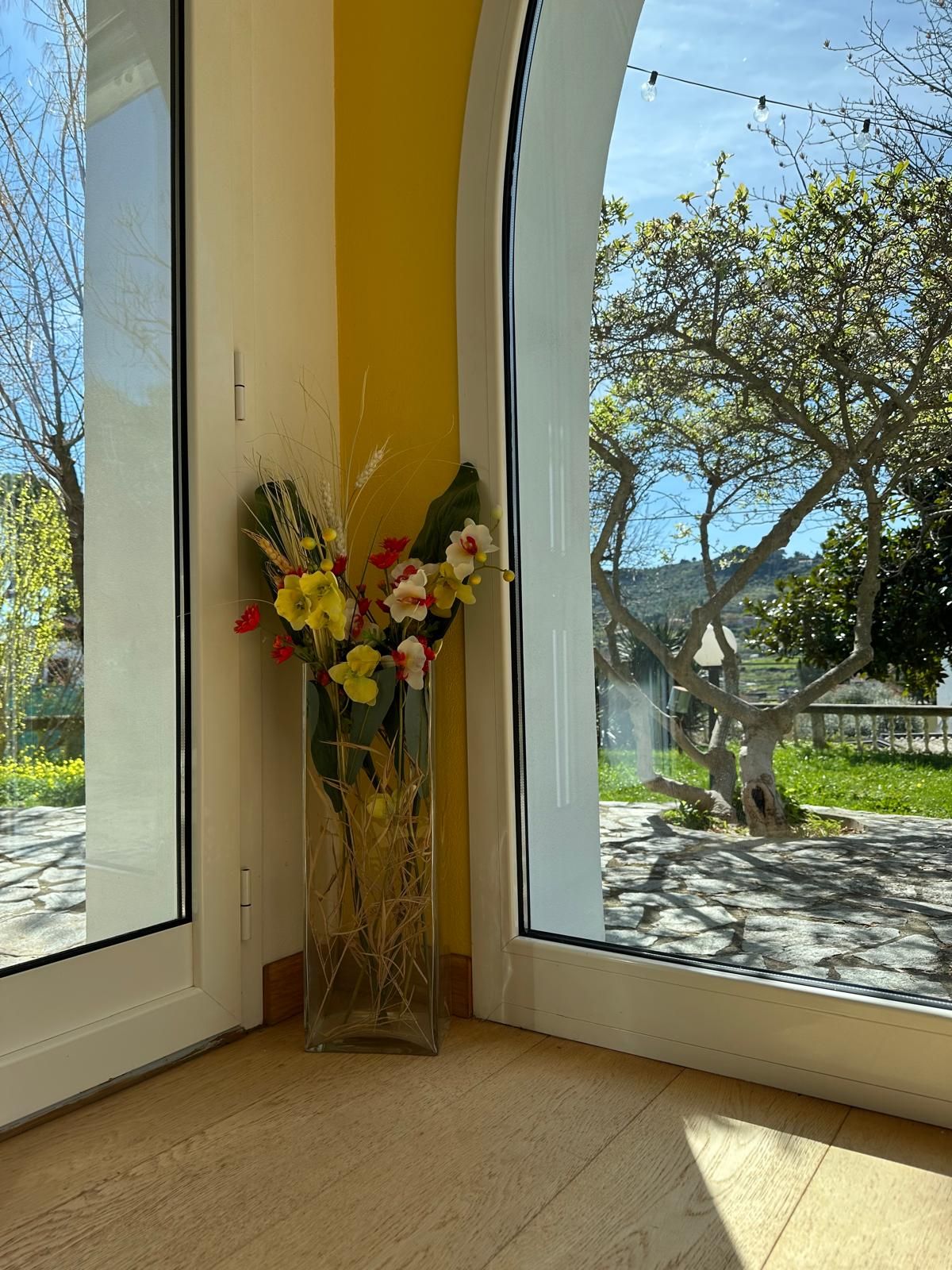 A room with a large window, a vase with flowers, and a view of trees and a pathway outside.