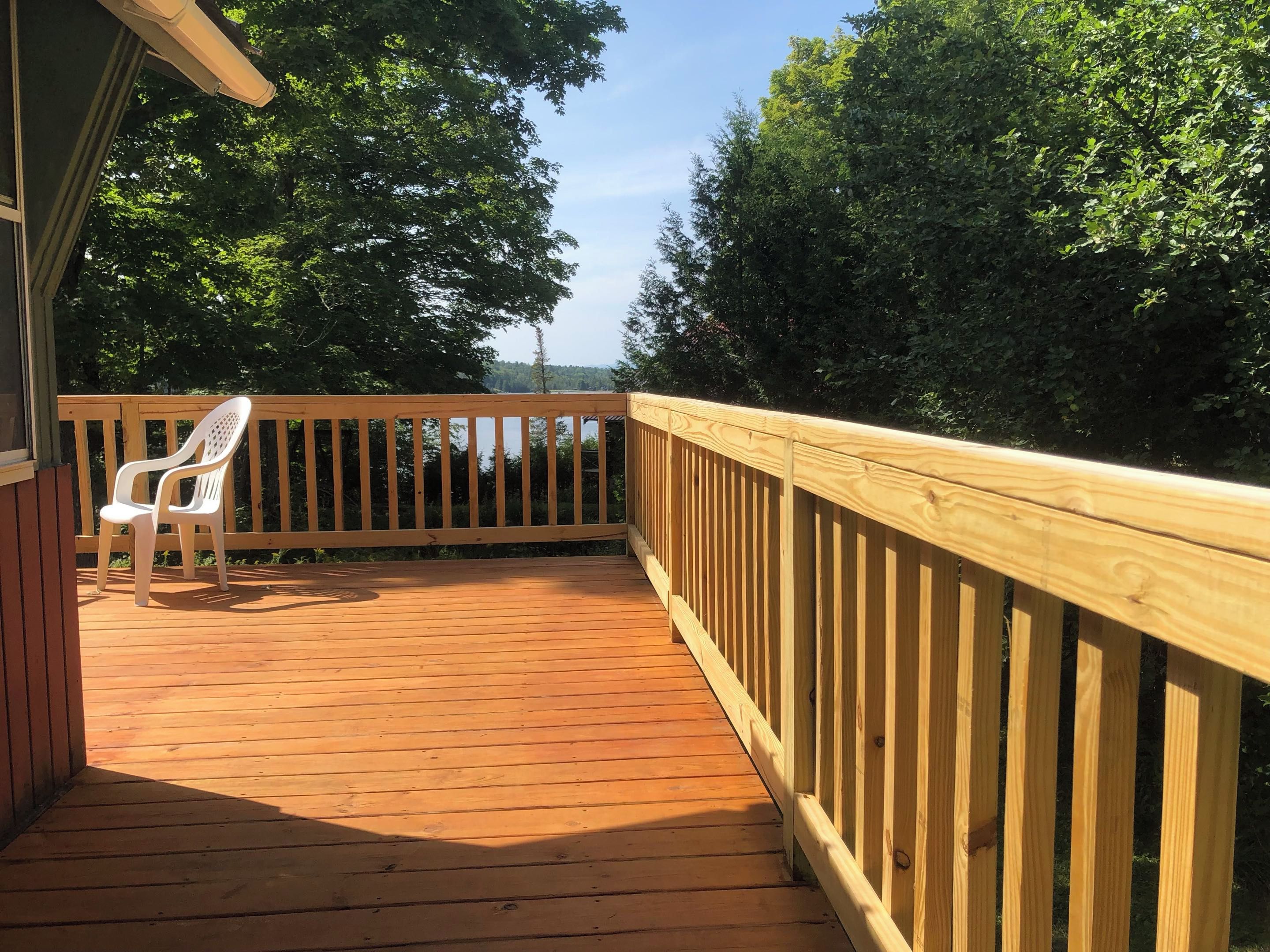 A wooden deck with a railing, a white plastic chair, and a view of a lake surrounded by trees.
