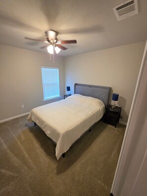 Une chambre à coucher comprenant un lit, un ventilateur de plafond, une fenêtre avec des stores et une table de chevet avec une lampe.