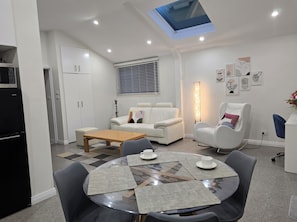 A modern living room with a sofa, a coffee table, and a dining area with a glass table and chairs. - New one of two 2-bedroom units in Gawler-Barossa. Unique garden. Close to shops (Evanston)