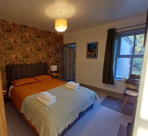 A bedroom with a large bed, a bedside table, a lamp, a window with curtains, and a door. - Stylishly Restored House in Oulton Village, between The Broads and The North Sea (Lowestoft)