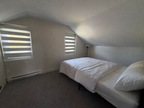 A bedroom with a bed, two windows with blinds, a radiator, and a ceiling light.