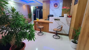 A hotel reception area with a marble counter, two bar stools, potted plants, and a logo on the wall.