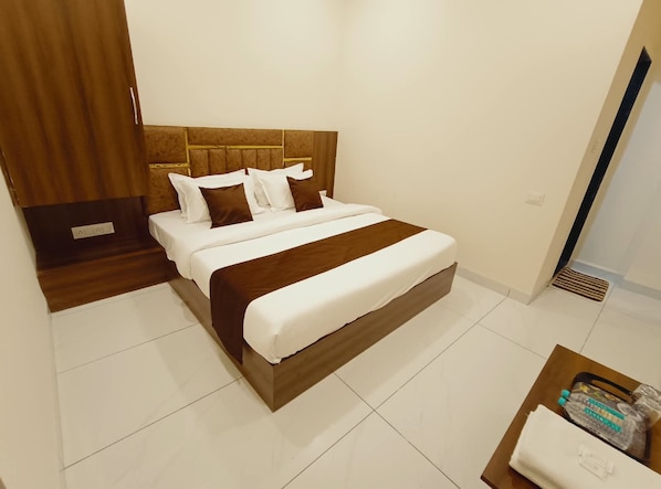 A hotel room with a bed, wooden headboard, white bedding, and a small table with a bag and a box. - Hotel Sai Kripa (Ujjain)