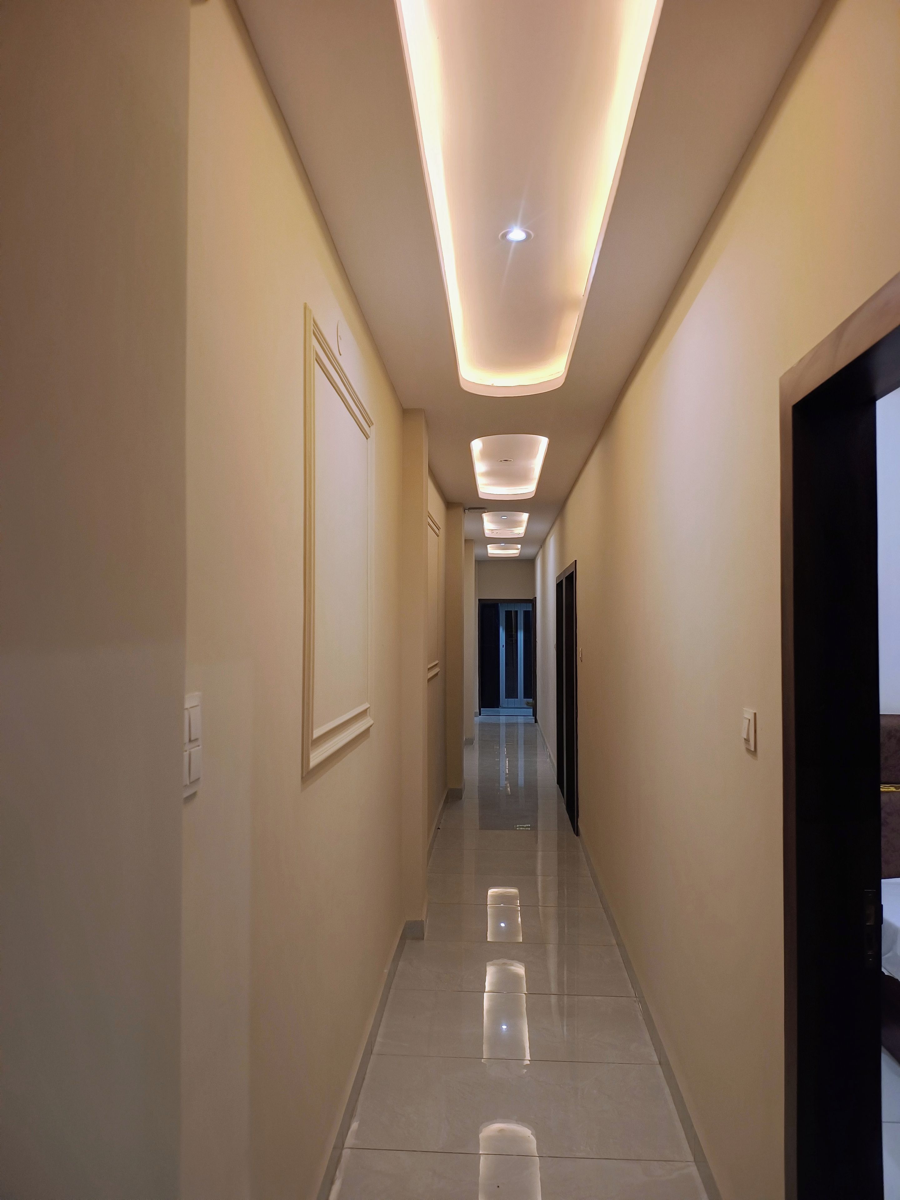Un pasillo de hotel con diseño moderno, que presenta un largo corredor con varios focos empotrados en el techo y un suelo de baldosas pulidas.