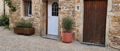 A stone building with a tiled roof, a white door, and a large wooden door. There are two potted plants in front.
