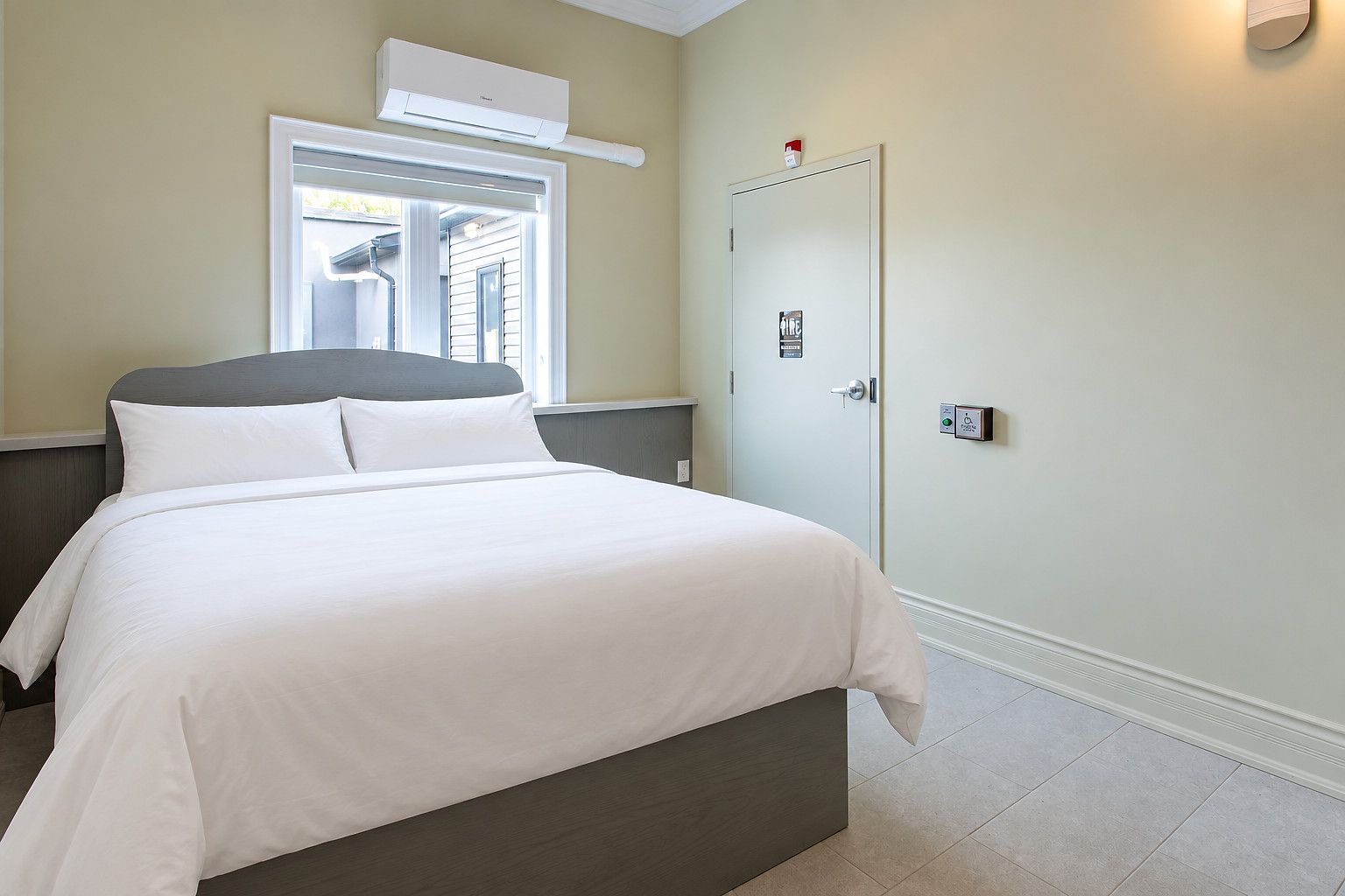 Ein Hotelzimmer mit Bett, Fenster, Klimaanlage, einer Tür mit einem Schild und einem Lichtschalter.