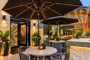 An outdoor seating area with round tables and umbrellas, a stone wall, and decorative plants. - Q W Hotels and Suites (Quinte West)
