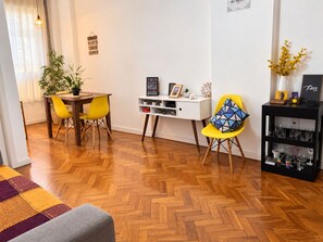 Ein modernes Wohnzimmer mit Holzdielen im Fischgrätmuster, einem Esstisch mit gelben Stühlen, einem weißen Konsolentisch mit Dekorationen und einem schwarzen Regal mit Gläsern.