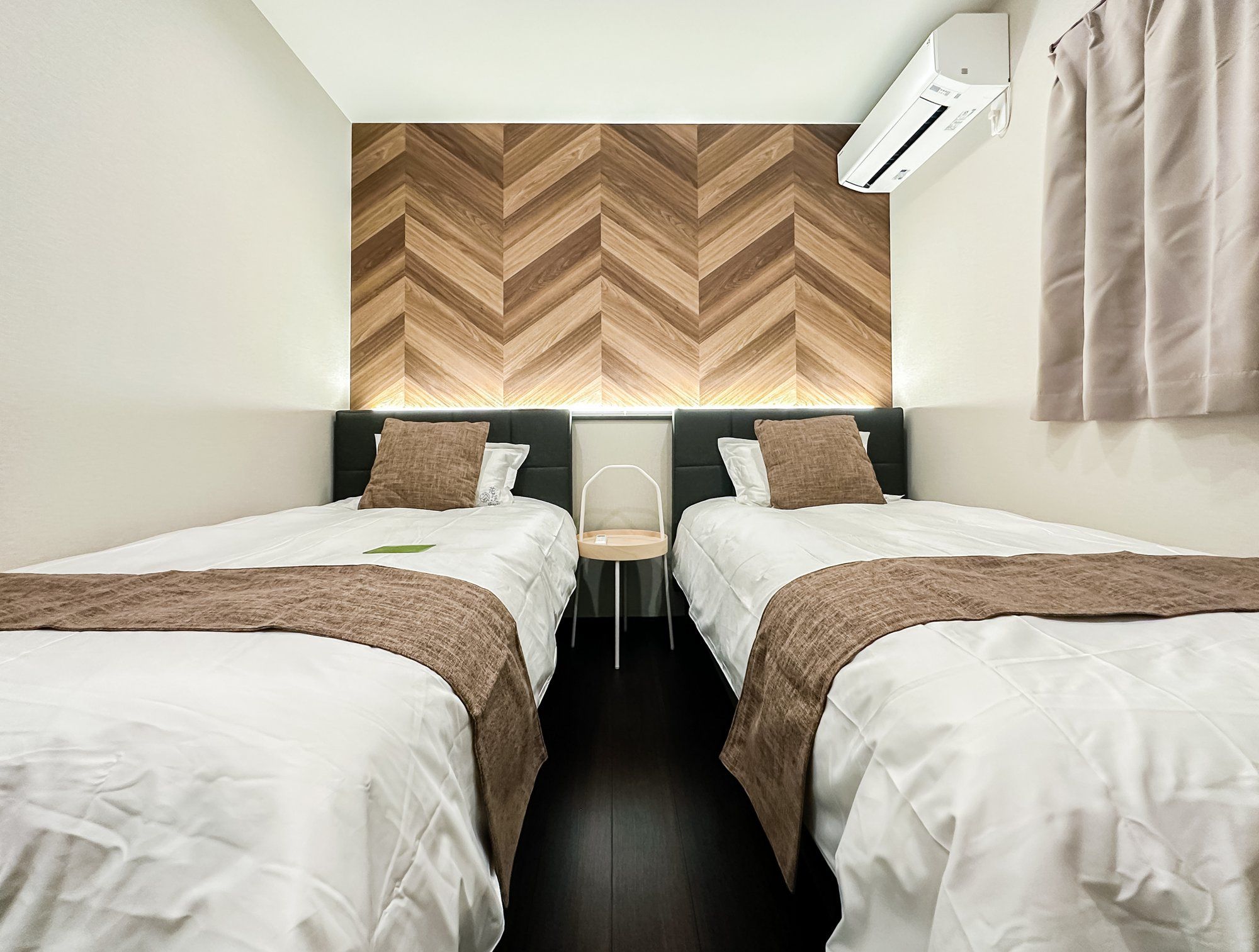 A hotel room with two beds, a wooden herringbone-patterned headboard, a small table, and an air conditioning unit.
