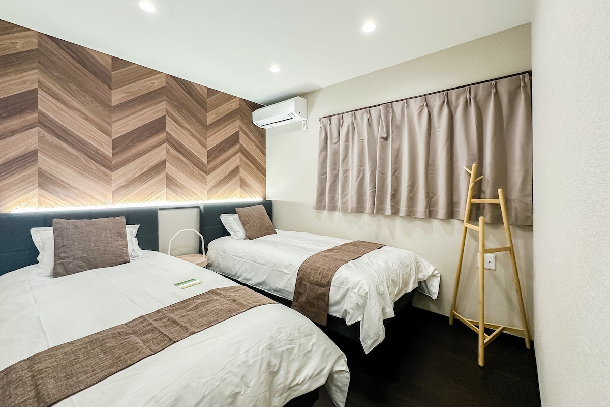 A hotel room with two beds, a wooden herringbone wall, an air conditioning unit, a window with curtains, and a wooden ladder.