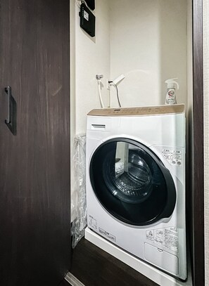 A compact, white front-loading washing machine with a wooden top, placed in a small laundry room with a wooden door and a wall-mounted electrical outlet. - HANASTAY KAERU (Osaka)