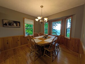 A dining room with a wooden table, chairs, a chandelier, and windows with a view of greenery. - 3-bedroom cabin in beautiful Ellijay with cool mountain vibes (Ellijay)