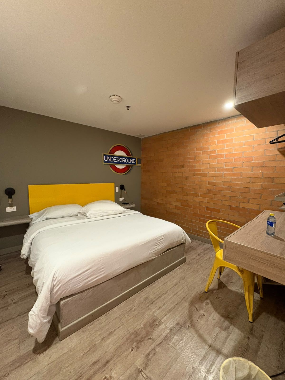 A hotel room with a bed, a table, a chair, a brick wall, and a sign indicating an underground station.