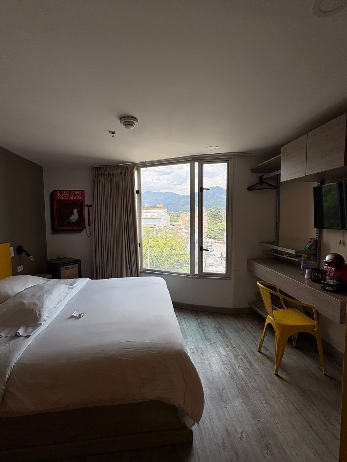 A hotel room with a bed, a desk, a chair, a window with a view of buildings and mountains, and a mounted television.